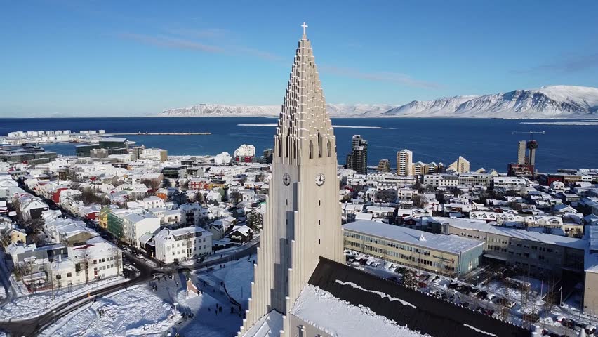 Aerial drone video soaring over snowy Reykjavík, capturing Hallgrimskirkja, white-covered rooftops, the bright blue sea, and mountains in the distance. Iceland’s winter wonderland. 