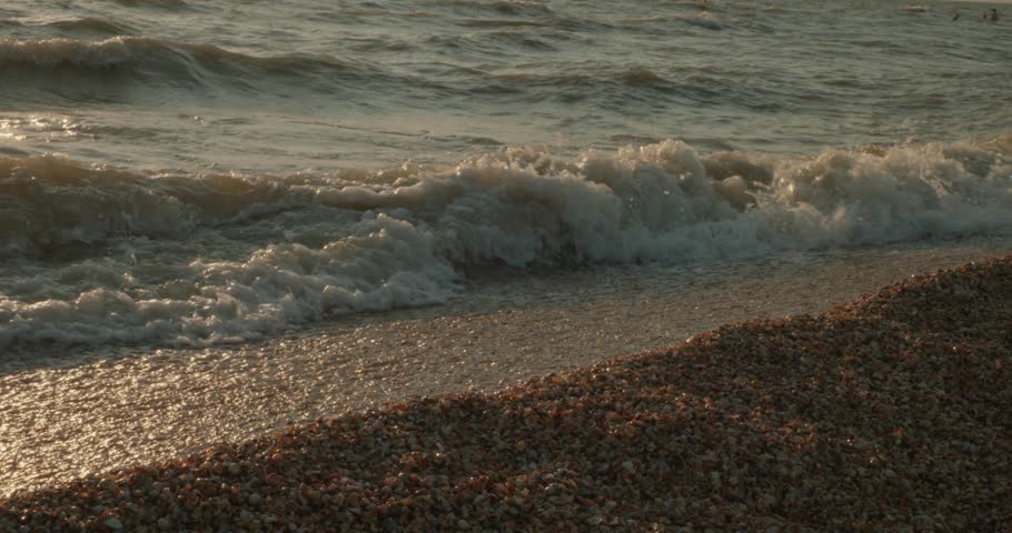 Golden sunset light reflecting on ocean waves as they gently crash and roll over a pebble beach, creating white foam along the serene coastline in a tranquil and beautiful natural scene.