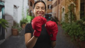 Woman holding red boxing gloves up in a narrow street beside an old building, smiling and sticking out tongue playfully; confidence empowerment. - Powered by Shutterstock - Get 15% off with code: PIKWIZARD15