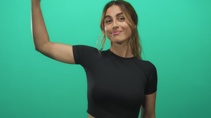 Young blonde woman wearing black crop top points finger to her flexed bicep in a studio; empowerment confidence.