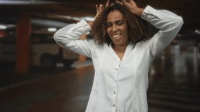 Woman smiling with hands on head, curly hair visible, wearing white shirt in indoor parking building among cars; joy. - Powered by Shutterstock - Get 15% off with code: PIKWIZARD15