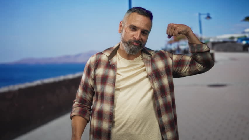 Man flexes bare arm on street promenade by the sea while looking at his bicep; strength confidence vitality.