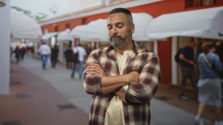 Man with crossed arms showing forearms on street lined with awnings and storefronts, wearing plaid shirt; confidence resilience strength.