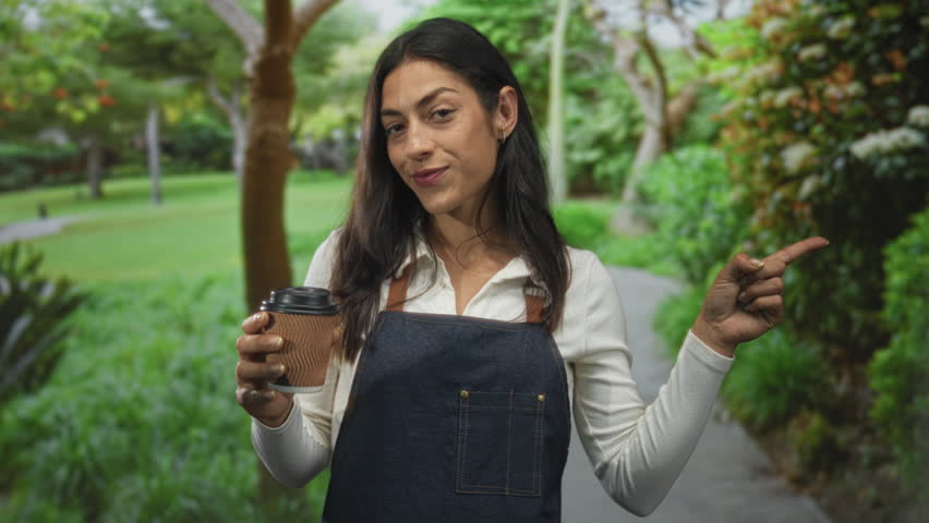 Woman in denim apron holding a takeaway coffee cup and pointing finger toward a park path in forest setting, smiling with visible hand and face; friendly service.