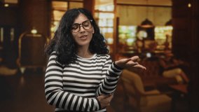 Woman brunette with glasses and open palm gesture wearing striped sweater in hotel lobby, presenting to camera; welcoming hospitality friendly conversation. - Powered by Shutterstock - Get 15% off with code: PIKWIZARD15