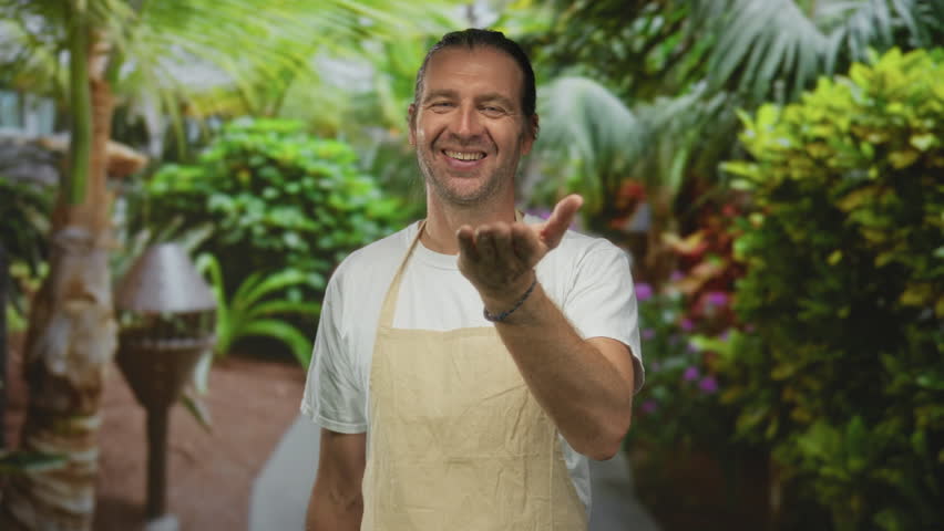 Man in beige apron blows a kiss with outstretched hand in green park setting; affection warmth connection joy.