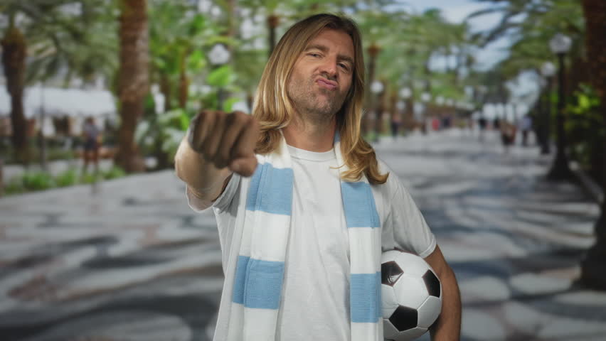 Blond man punching with fist while holding a soccer ball on a bustling street lined with palm trees; passion.