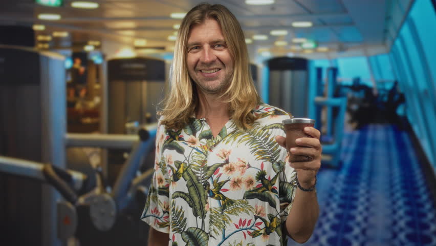 Man with long blond hair wearing a floral shirt smiling while holding cup in a bright studio filled with gym equipment; contentment.