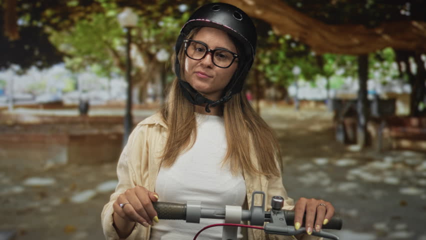 Young woman on electric scooter points finger to helmet while holding handlebar on street lined with trees and benches; playful curiosity.