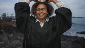 Woman judge in black robe smiling with hands forming a heart over head, glasses visible, posed against a seaside studio set with rocks; pride achievement. - Powered by Shutterstock - Get 15% off with code: PIKWIZARD15