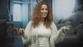 Young woman holding smartphone with raised hand and broad smile in building foyer, hand visible and curly hair; joyful confidence. - Powered by Shutterstock - Get 15% off with code: PIKWIZARD15