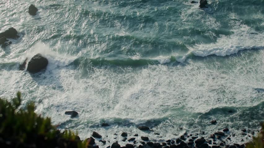 Foamy waves crash against a rocky seashore from a top-down view, revealing textured rocks, shallow waters, and dynamic coastal movement in a natural scenic setting.