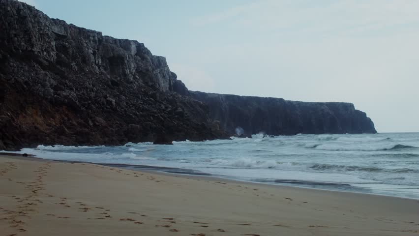 Foamy waves crash against the sandy shore at the base of steep cliffs during dusk, creating a dramatic coastal scene with soft light and natural ocean movement.