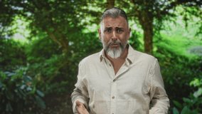 Man with grey beard and open linen shirt standing among trees in forest park, hand extended forward and mouth open as if speaking; concern pleading. - Powered by Shutterstock - Get 15% off with code: PIKWIZARD15