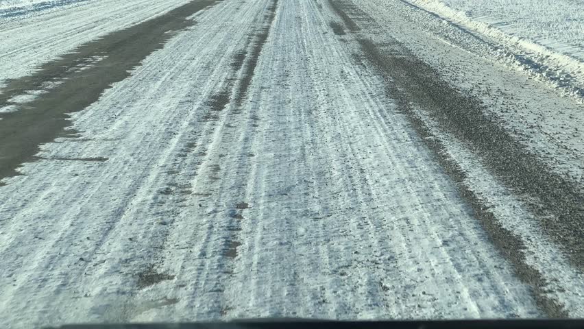 Sparse rural roadway coated with frost and snow, continuing straight across farm fields and prairie soil, illustrating cold-distance travel through winter countryside. Siksika Alberta Canada.