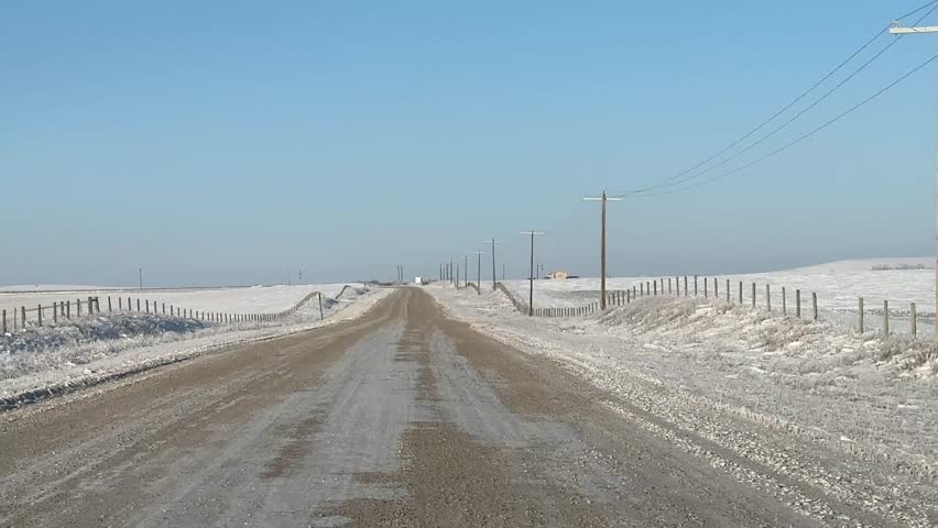 Long isolated winter roadway stretching across open prairies, covered in packed snow and ice, leading toward a distant horizon under a pale sky and surrounded by quiet farmland.