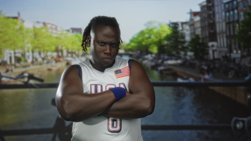 Man with arms crossed on street by amsterdam canal wearing usa jersey and wristband; defiance strength pride.