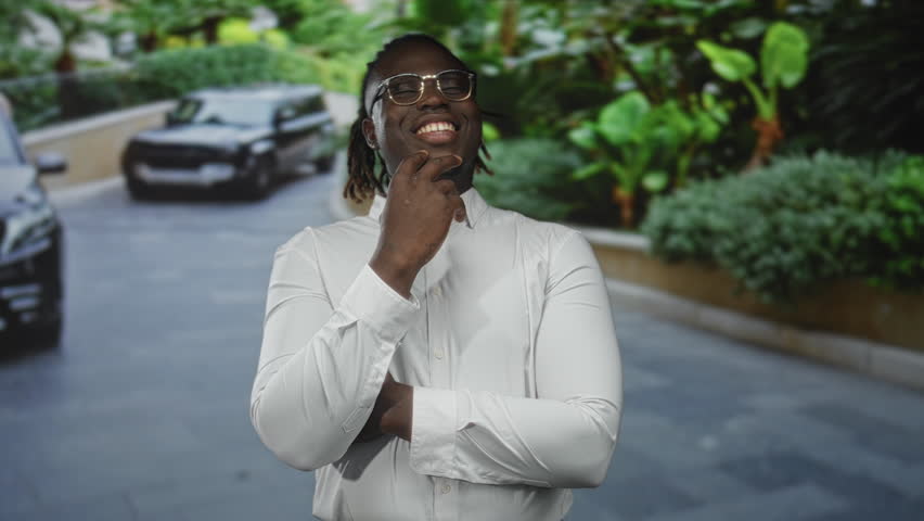 Man with hand on chin smiling, wearing glasses and white shirt on street driveway with cars and plants; confidence aspiration.