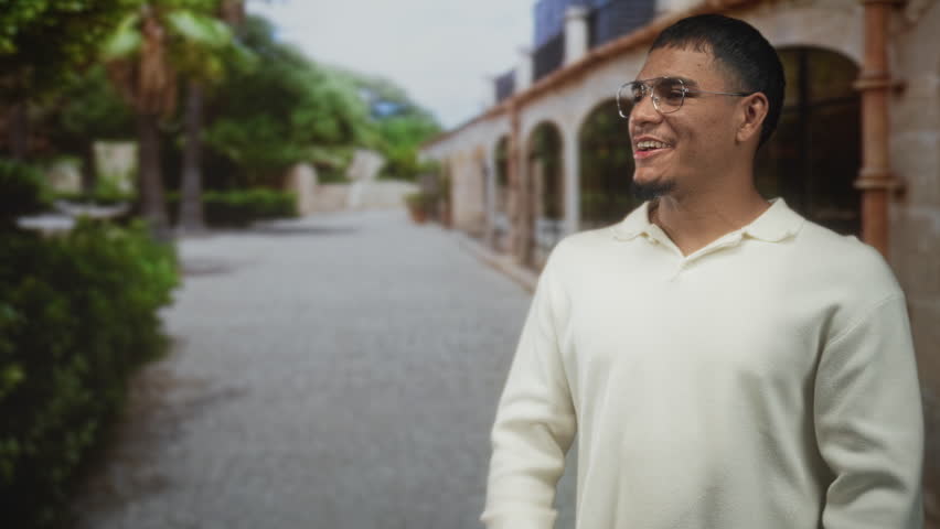 Man clapping hands on a cobbled street beside an arched building and palm trees, wearing glasses and cream sweater; celebration joy.