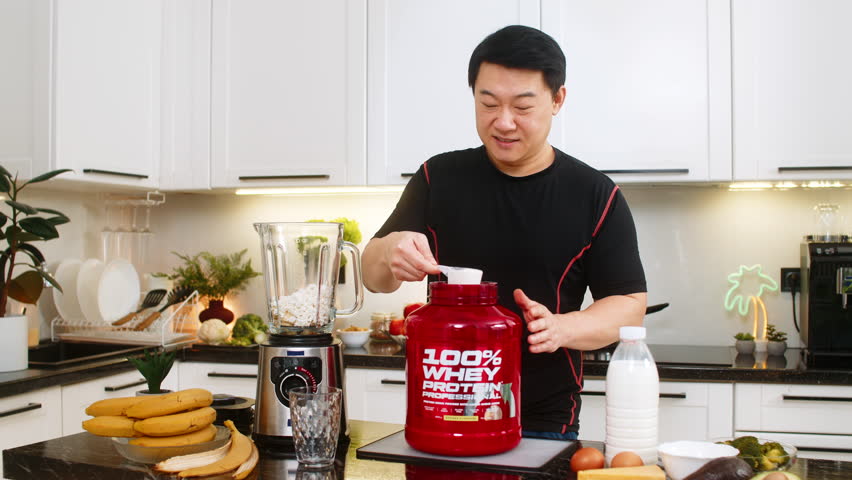 Asian young adult man pours whey protein into blender with banana and cottage cheese preparing shake. Protein smoothie supports muscle gain weight control fitness energy and home kitchen healthy diet.