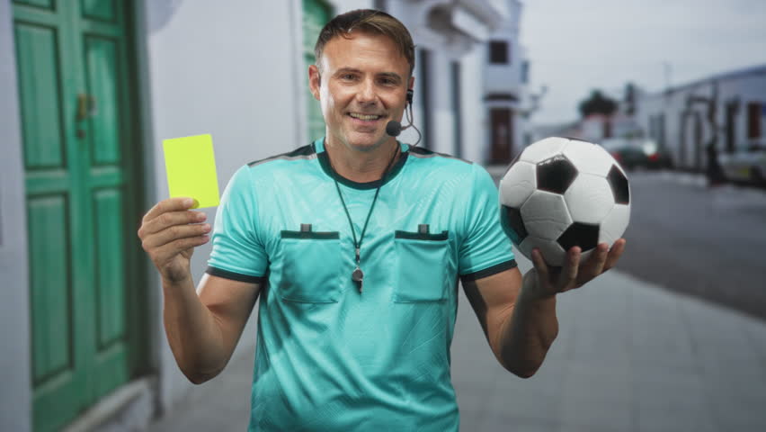 Man referee holds yellow card in left hand and soccer ball in right, whistle on lanyard and headset visible on a street; authority fairness.