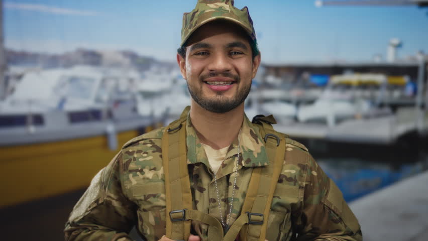 Man in camouflage uniform smiles and holds backpack strap showing dogtags with boat in view at port; pride duty.