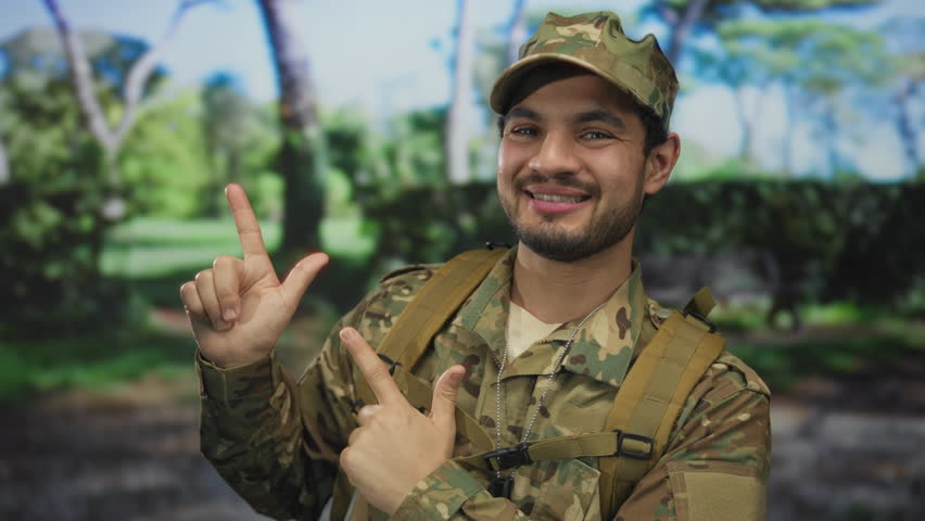 Man in digital camouflage uniform with tactical vest and backpack finger pointing to object in green forest scene under sunlight; pride duty.