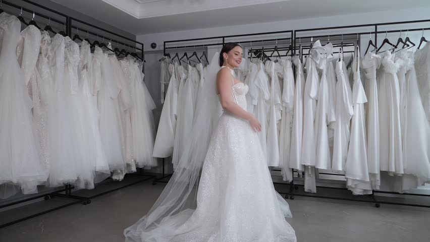 A graceful bride in slow motion, her long veil swirling, stands in the wedding salon, surrounded by numerous white dresses hanging on a rack. A woman tries on a wedding gown and twirls.