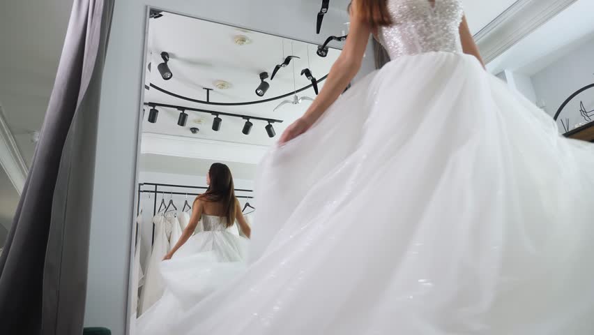 A happy woman is trying on a wedding dress in a wedding salon. She is standing in front of the mirror and spinning, and the long chiffon skirt of her wedding dress flutters in different directions.