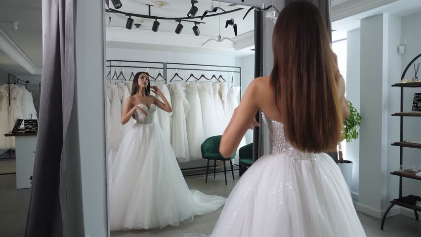 A happy woman is trying on a wedding dress in a wedding salon. She is standing in front of the mirror and spinning, and the long chiffon skirt of her wedding dress flutters in different directions.