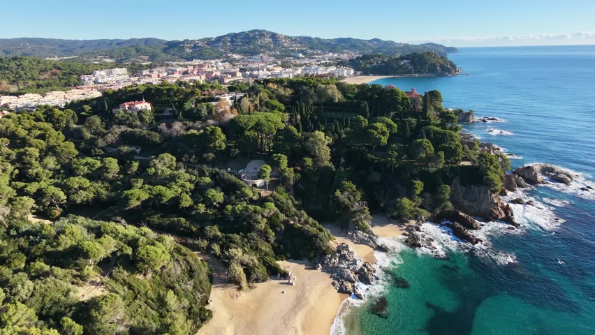 A smooth and cinematic drone tracking shot gracefully moves parallel to the shoreline of Lloret de Mar, highlighting the contrast between the deep blue Mediterranean Sea and the characteristic pine
