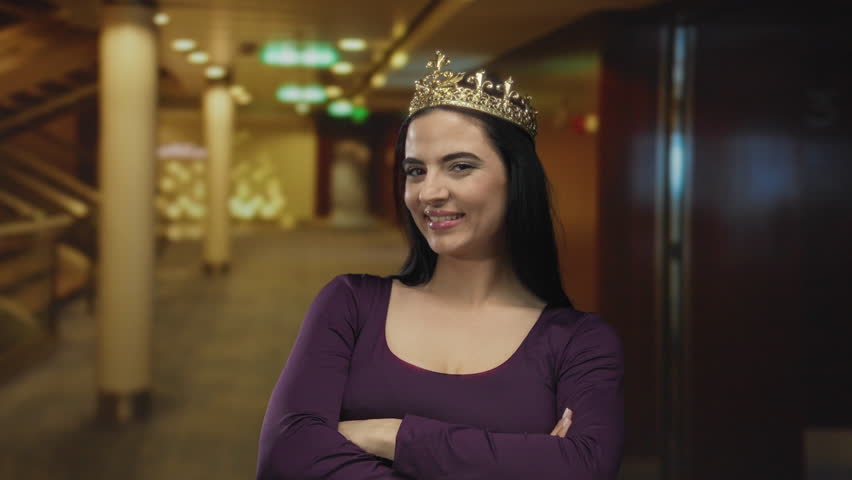 Woman smiling confidently indoors wearing a crown and purple dress in a vibrant setting with columns and warm lights.