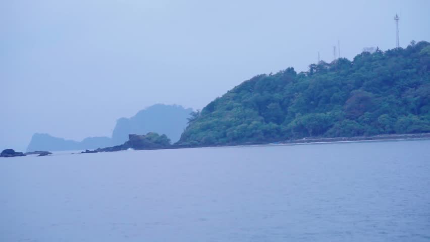 A beautiful, serene shot of a tropical island shoreline with lush green jungle meeting the calm blue sea under a soft, overcast sky, viewed from a moving boat