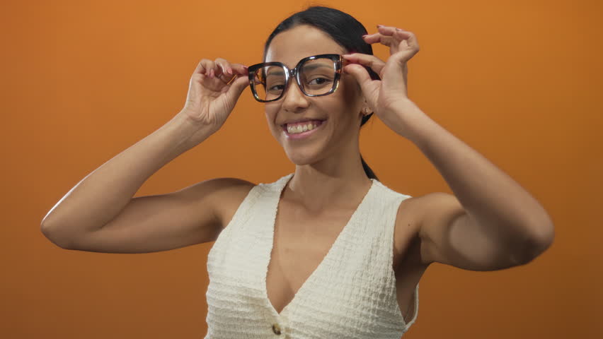 Young hispanic woman smiling confidently with glasses and a thumbs up gesture against an isolated orange background wall showcasing positivity and approval.