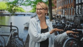 Woman with short blonde hair and glasses presenting open palms beside parked bicycles on a canal street near brick buildings; joy friendly invitation. - Powered by Shutterstock - Get 15% off with code: PIKWIZARD15