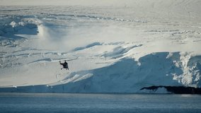 Red helicopter skims past towering ice cliffs along a frozen coastline, dwarfed by blue glacier walls and stratified snowfields above calm polar waters during an aerial traverse. - Powered by Shutterstock - Get 15% off with code: PIKWIZARD15