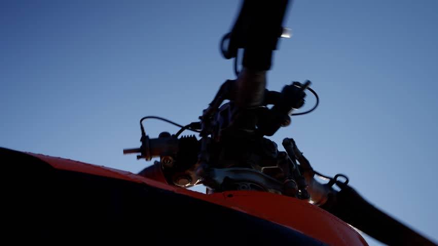 Close-up view of a helicopter rotor hub and swashplate assembly with blades silhouetted against a blue sky, emphasizing mechanical detail and precision in aviation engineering