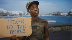 Woman in camouflage uniform holds protest sign and points finger to sign at seaside port; empowerment. - Powered by Shutterstock - Get 15% off with code: PIKWIZARD15