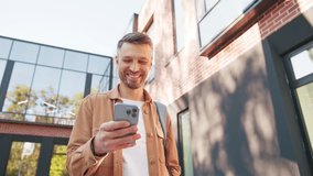 Attractive Caucasian male holding smartphone and smiling while enjoying lighthearted video content. Man carrying backpack standing near modern brick facade. Cheerful male taking brief pause. - Powered by Shutterstock - Get 15% off with code: PIKWIZARD15