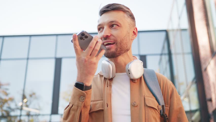 Caucasian male holding smartphone horizontally and recording voice message. Man wearing headphones around neck standing near bright glass facade. Focused male sharing quick audio update.