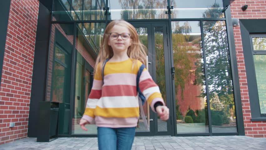 Caucasian schoolgirl wearing glasses running toward camera along paved walkway beside brick building. Child carrying backpack and smiling while moving energetically after leaving school.
