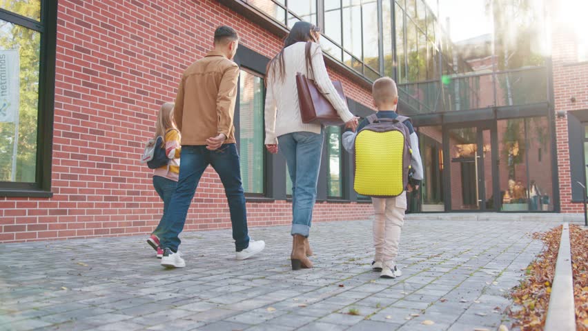 Four family members walking toward school walkway. Parents guiding boy with bright backpack and girl with colorful sweater. Stepping along brick path morning routine. Supporting children education.