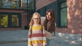 Schoolgirl standing smiling toward camera on brick walkway. Child wearing glasses carrying backpack while waiting. Female parent behind adjusting straps preparing daughter for walk home. - Powered by Shutterstock - Get 15% off with code: PIKWIZARD15