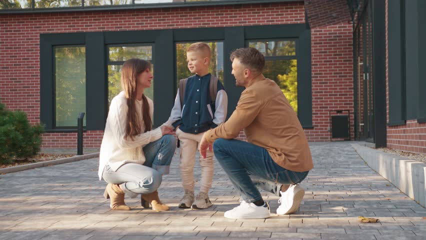 Caucasian parents kneeling and talking with son near school building. Mother checking his jacket while speaking. Father watching and encouraging child during morning routine before heading to class.