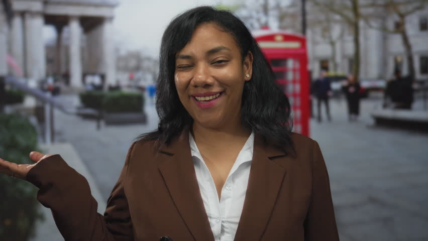 Woman extends hand palm up on busy city street with smiling face and red phone booth visible; optimism.