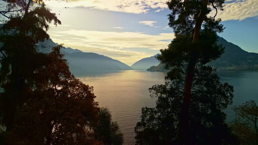 Lugano, Switzerland – December 2, 2024: Sunlit view from Parco Ciani over Lake Lugano toward Monte San Salvatore, Monte Caprino, and Paradiso, framed by cypress, larch, and pine trees.