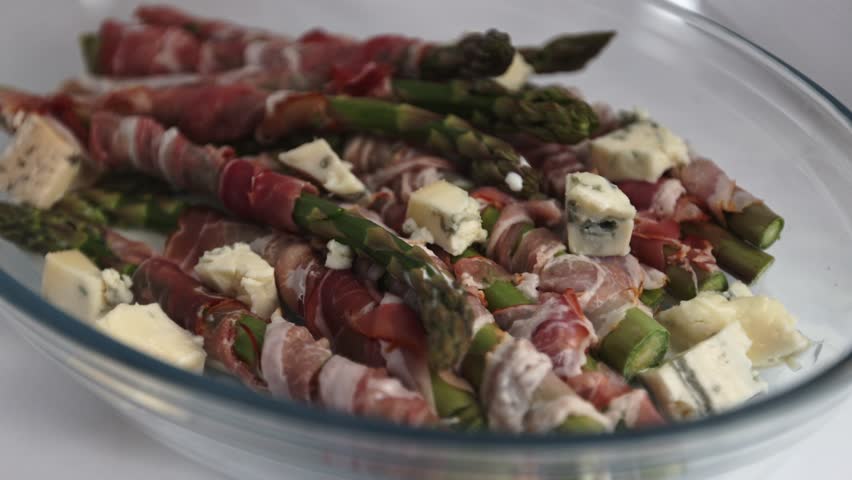 A clear baking dish filled with green asparagus wrapped in bacon and sprinkled with diced gorgonzola, photographed against a smooth white background.