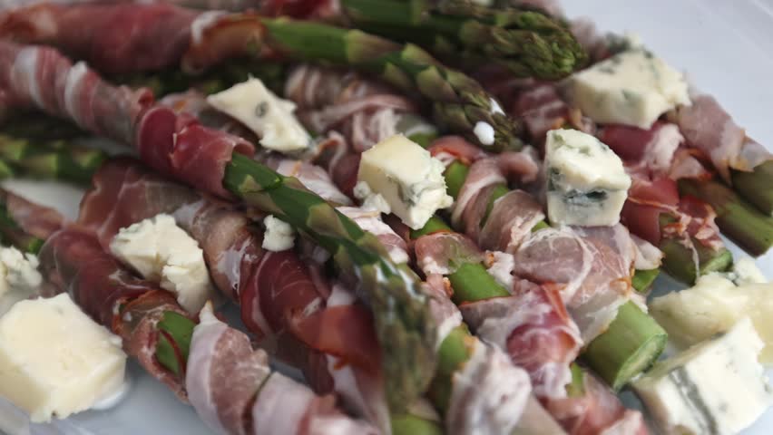 Crispy bacon-wrapped asparagus arranged neatly in a transparent oven-safe dish, with gorgonzola pieces filling the gaps, displayed on a bright white surface.