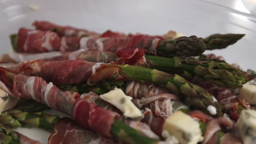 Fresh asparagus bundles coated in bacon rest in a glass oven tray, surrounded by evenly scattered gorgonzola cubes, captured on a minimal white background.