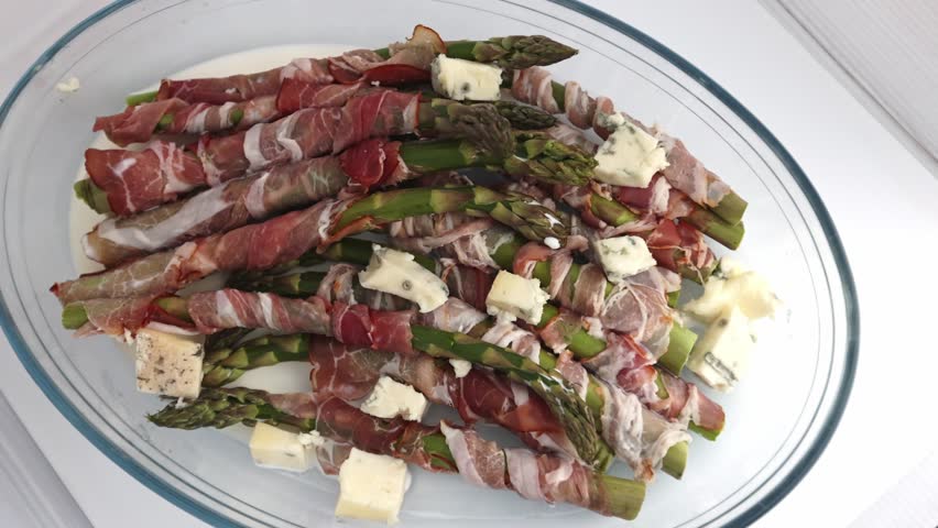 Bacon-wrapped asparagus spears laid in a clear baking dish, complemented by small cubes of gorgonzola nestled between them, shot on a clean white backdrop.